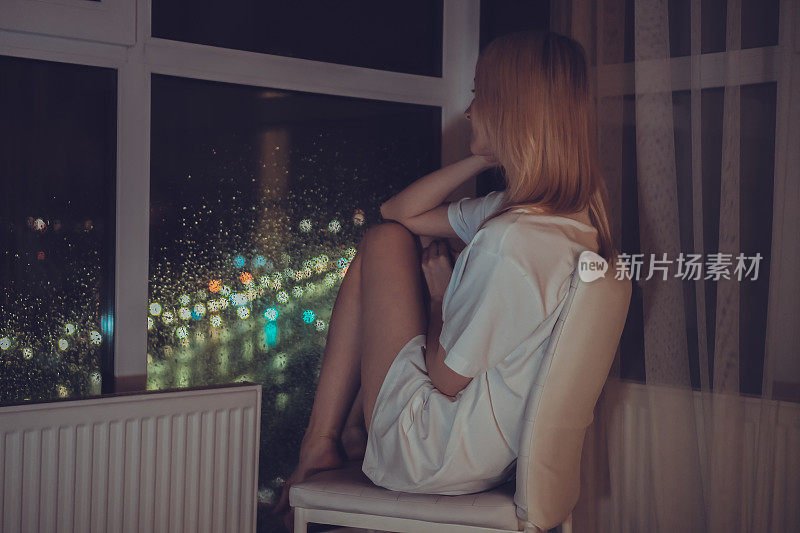 穿着白衬衫的金发女郎坐在椅子上，下巴托着，透过落地窗望着外面的夜城