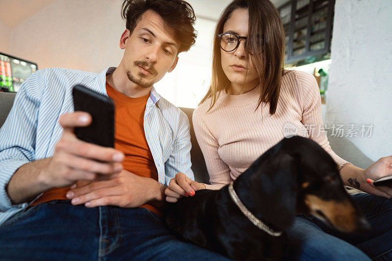 一对年轻夫妇在家一边玩智能手机一边和他们的狗狗放松