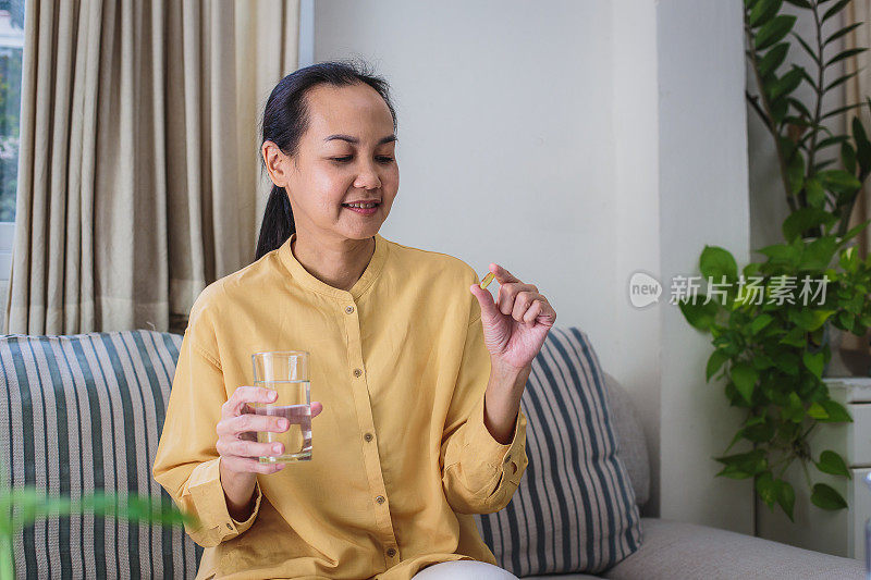 亚洲成熟女性坐在家里的客厅里，一边吃药或服用补充剂，一边手持水杯