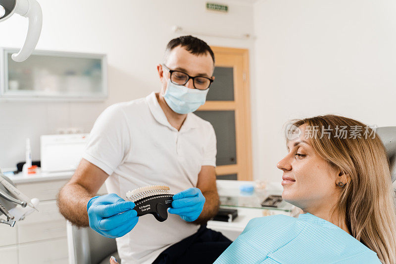 妇女看着贴面或种植牙配色样本在医生手中。牙医。牙科医生在牙科诊所为女性病人示范牙齿美白的颜色深浅。