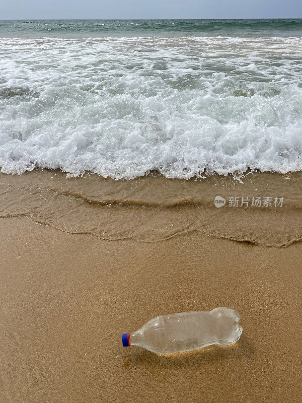 图像的空，透明的塑料饮料瓶与蓝色盖子被冲到沙滩在退潮水的边缘，破碎的海浪冲刷海洋垃圾和污染的海岸，垃圾的沙子，肮脏的海滩，海洋背景，重点在前景