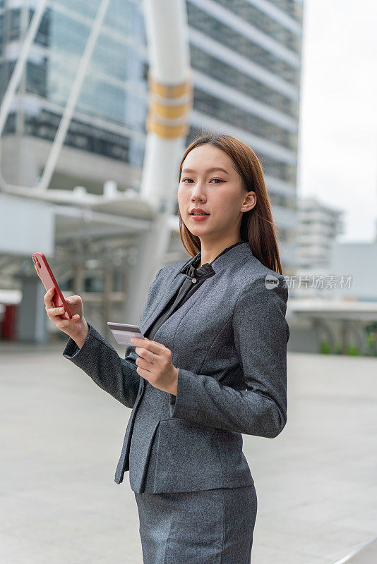 一名年轻的亚洲女商人穿着灰色的办公套装，在商业区用她的智能手机使用信用卡进行在线交易，她微笑着自信