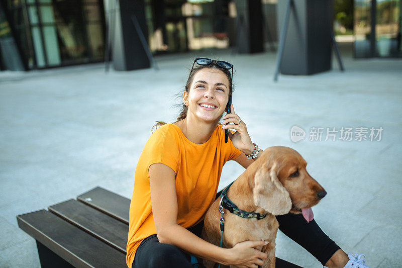 年轻女子和她的可卡犬坐在长凳上讲电话