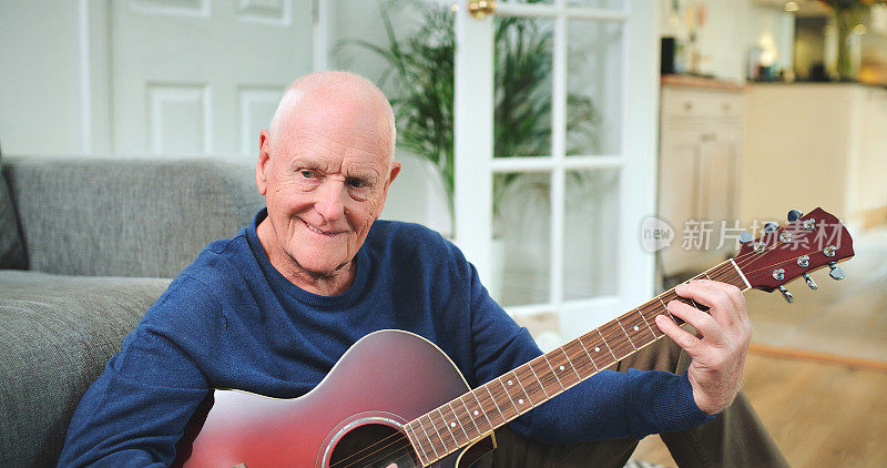 老人拿着吉他，微笑着在家里的客厅里演奏音乐。资深人士，原声乐器和快乐的男音乐家在休息室为声音，音频和创作人才表演