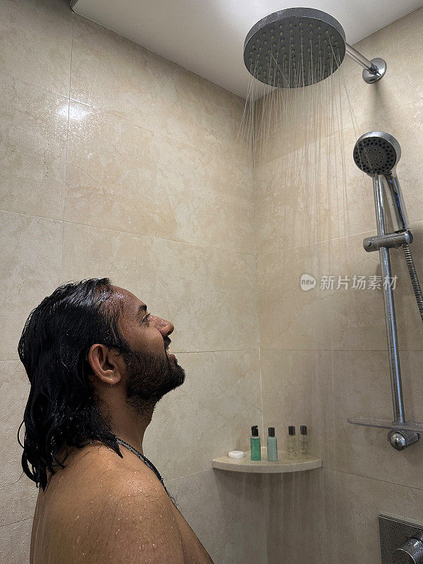 一名印度男子在淋浴间洗头，洗发水泡沫被流水冲洗掉，他抬头看，架子上的洗漱瓶，棕色马赛克瓷砖的墙壁，侧面视图，聚焦于前景