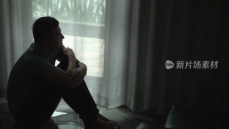 亚洲男人的剪影独自坐在黑暗的房间窗边，双手抱着头，感觉悲伤，沮丧，绝望，精神困扰。