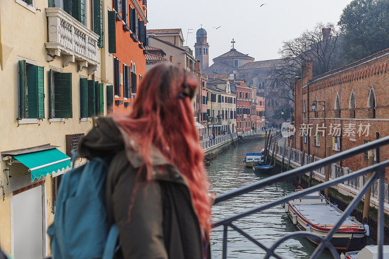 意大利威尼斯，一个染红头发背着双肩包的年轻女子站在运河上的一座桥上
