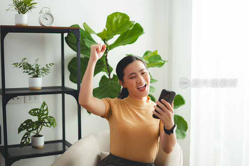 亚洲女性坐在家里的扶手椅上，用智能手机开心兴奋地做出了获胜的手势。