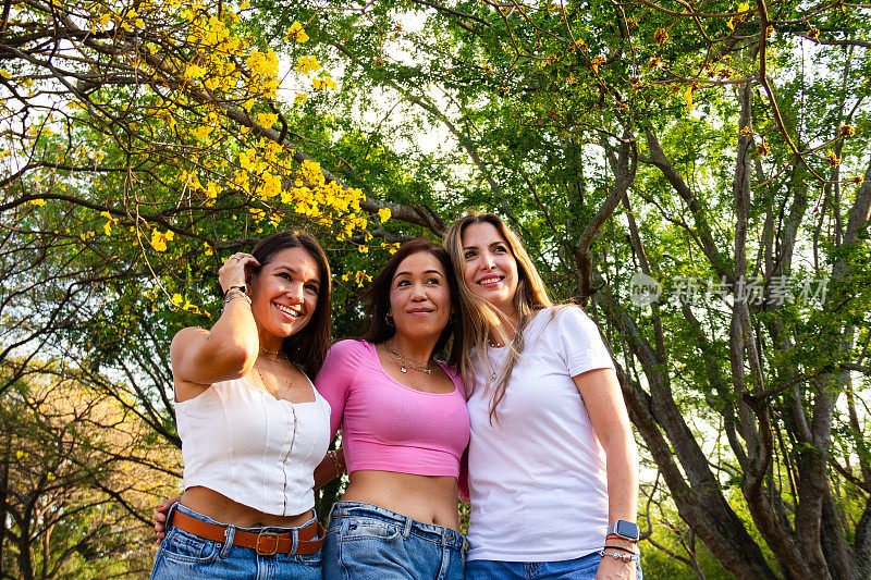 三位微笑的拉丁女性朋友在大自然中摆姿势拍照，目光转向另一个方向