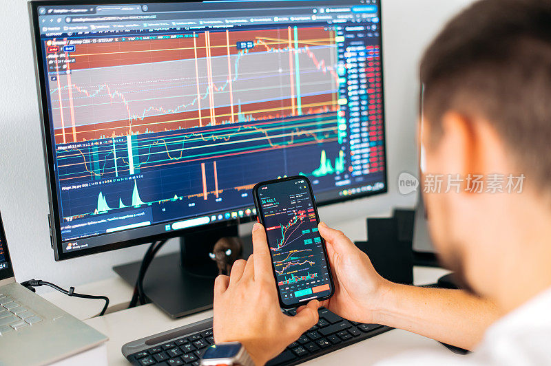 男性手中的智能手机，屏幕上显示交易数据指数图表，手机应用程序分析加密货币市场。加密货币交易员坐在他的工作场所前的显示器与图表