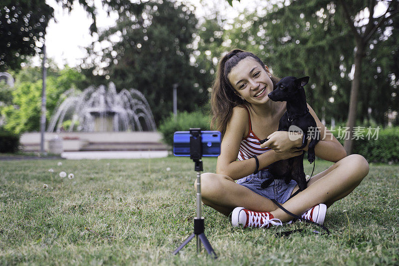 美丽的少女微笑着用手机在公园里拍宠物狗视频。网红录制视频。