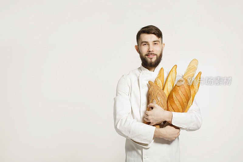 英俊微笑的面包师在制服拿着法棍面包架在白色的背景。英俊的男人拿着温暖的面包在他的手在白色的背景。