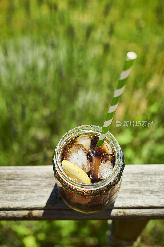 在夏日阳光明媚的花园里，一个盛满可乐、冰块和一片柠檬的玻璃瓶。