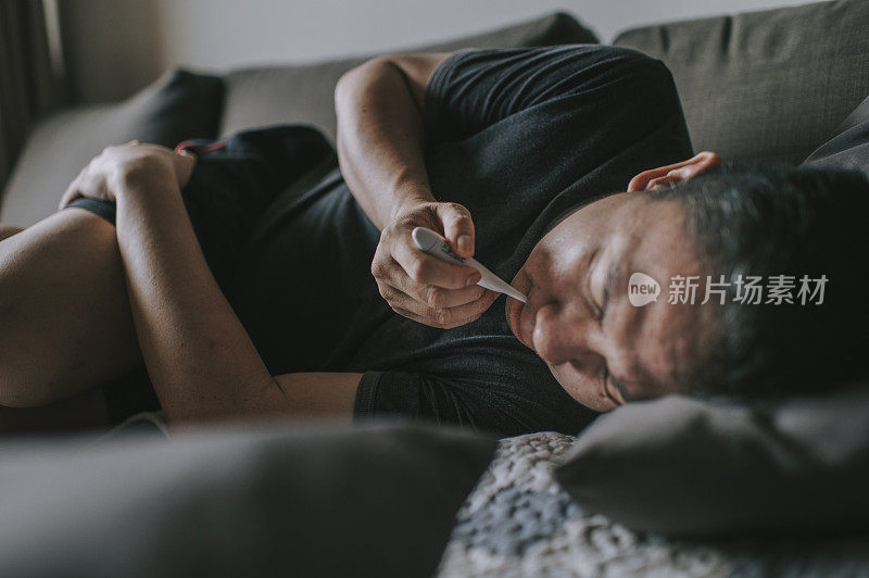 一名亚裔华人中年男子在客厅躺在沙发上休息，带着流感症状用体温计测量体温