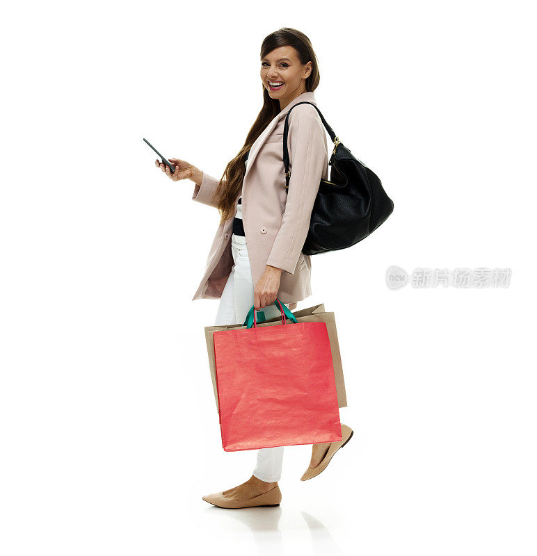 白人年轻女性穿着运动夹克，拿着钱包，使用智能手机走在白色背景前