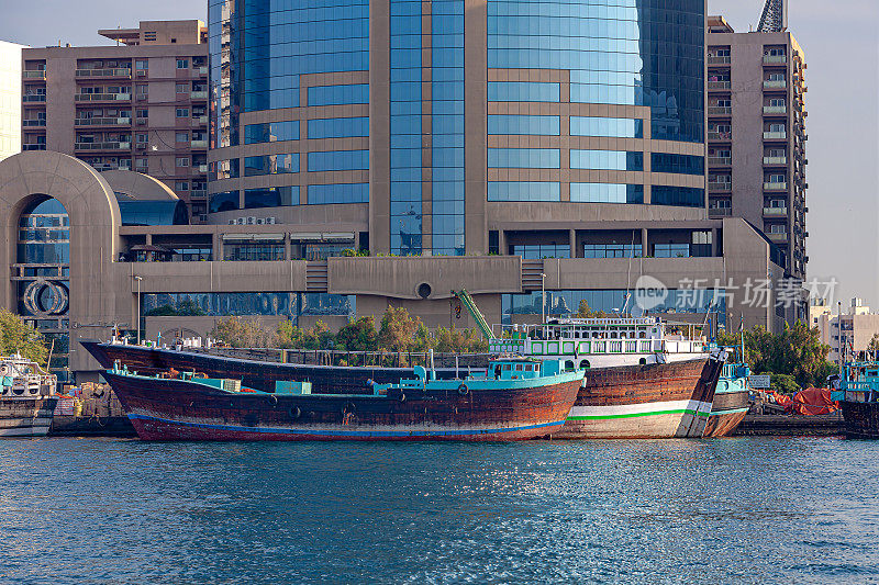 阿拉伯联合酋长国迪拜——早晨的阳光下，三艘传统的阿拉伯单桅帆船并排停泊在迪拜河德拉一侧的码头上;背景中，现代办公建筑。