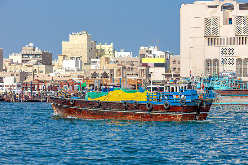 阿拉伯联合酋长国迪拜——一艘满载传统阿拉伯单桅帆船在晨曦中驶出迪拜河;在香料市场附近的Deira侧和Abra站，单桅帆船并排停泊;在遥远的背景，老