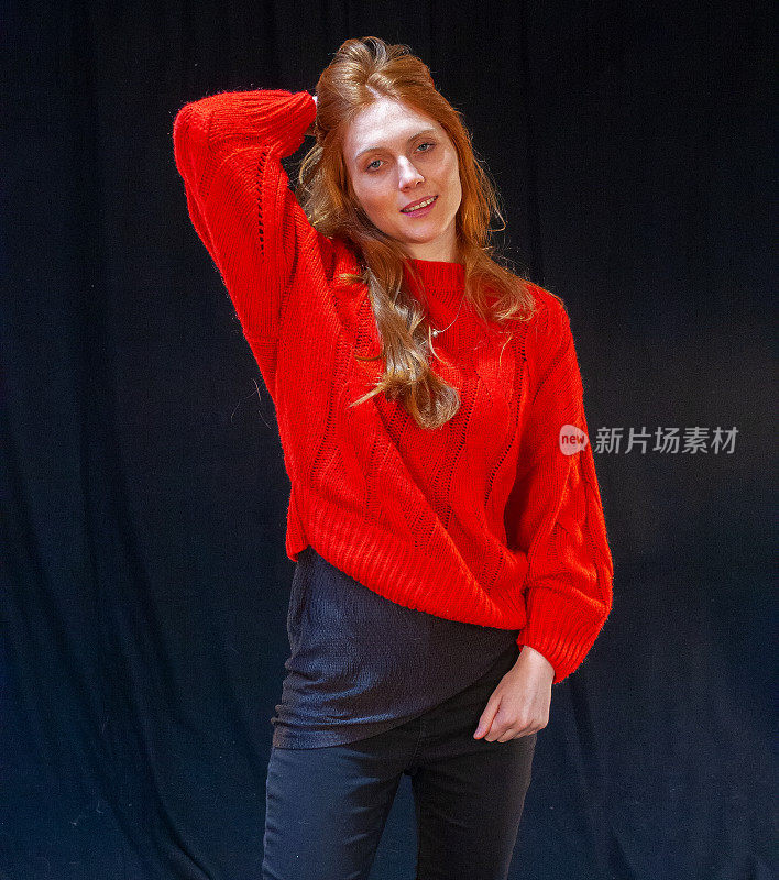 手挽着头发，一名年轻女子穿着一件红色毛衣，摆着放松的姿势，露出她长长的红色鬈发