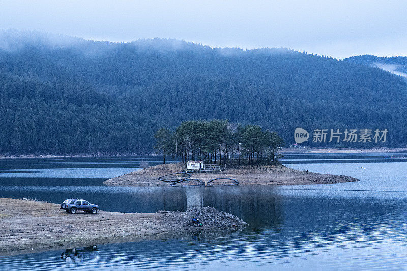 在雾中钓鱼。一辆停在湖岸的秋景车，和一个在水上小岛上移动的家。