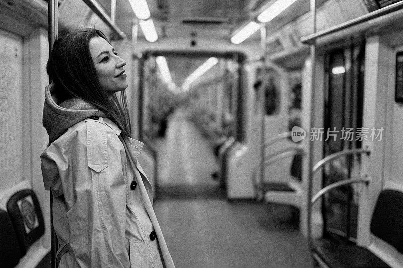 一个穿着米色风衣的女孩坐在地铁车厢里。BW