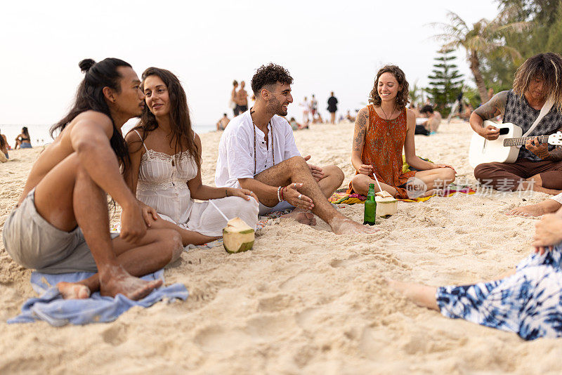 一群多种族的朋友坐在沙滩上，唱歌，弹吉他