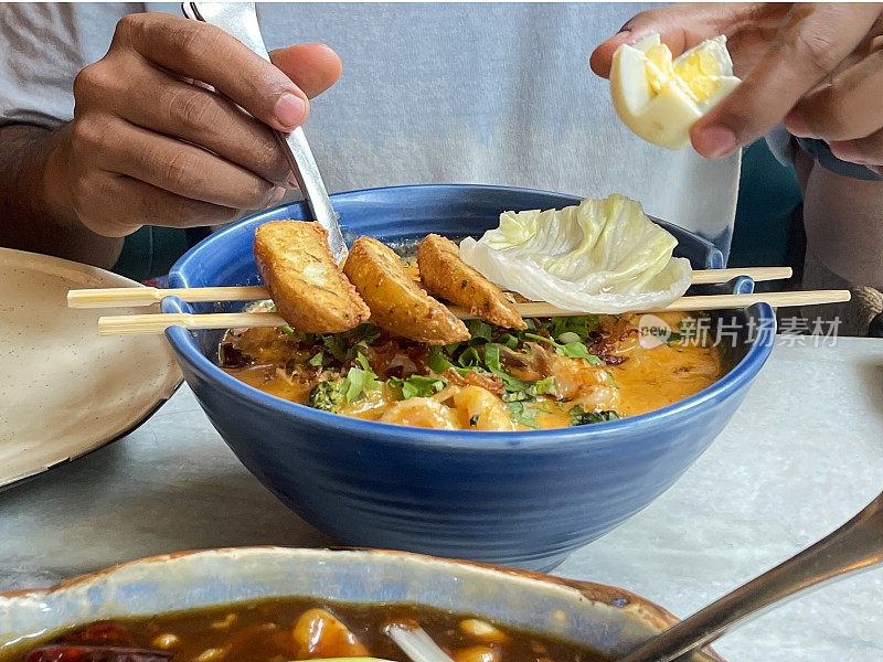 木筷子搁在自制的刻字碗上，辣椰奶叻沙汤配大虾，面目全非的人拿着煮熟的鸡蛋，生菜叶子和土豆楔子放在筷子上，金属勺子，重点放在前景