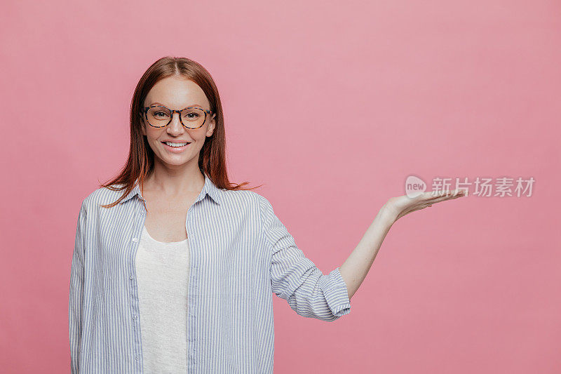 积极微笑的女人穿着优雅，举起手掌，拿着复印空间，戴着眼镜和衬衫，孤立在粉红色的工作室墙壁上，用高兴的表情做广告。促销的概念