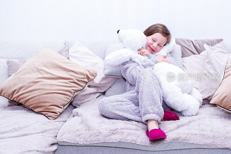 一幅白人儿童的肖像，一个7岁的女孩穿着长毛绒工作服，坐在柔软的大沙发上，抱着一个柔软的玩具——北极熊。