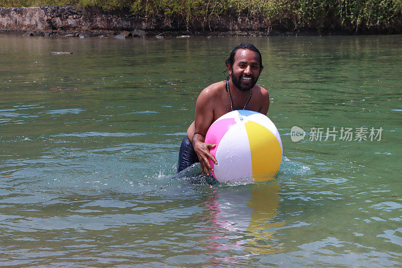 特写:一名穿着冲浪短裤的印度男子站在齐腰深的泻湖平静的水中，手里拿着漂浮的彩色塑料沙滩球，热带岛屿天堂，林地和棕榈树的景色，重点放在前景上