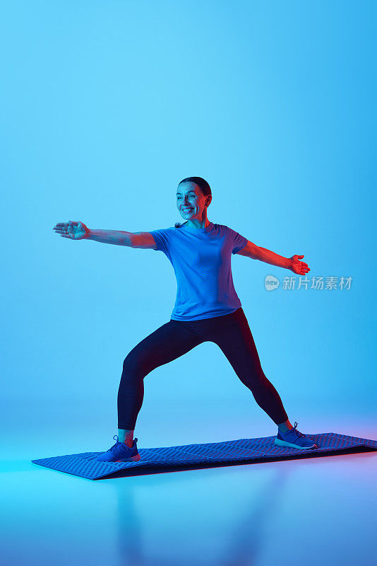 瑜伽和伸展运动。积极成熟的女人站在健身matt和训练的蓝色工作室背景在霓虹灯