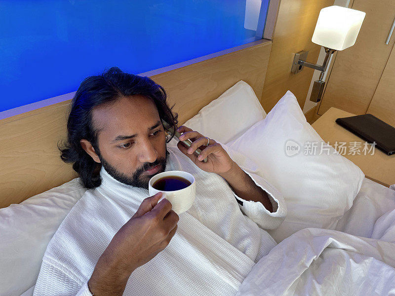 特写照片:印度男子躺在双人床上，穿着白色毛巾浴袍，躺在堆叠的枕头上，蜷缩在白色羽绒被下，一边从杯子里喝黑咖啡，一边用智能手机打电话，抬高视角，聚焦于前景