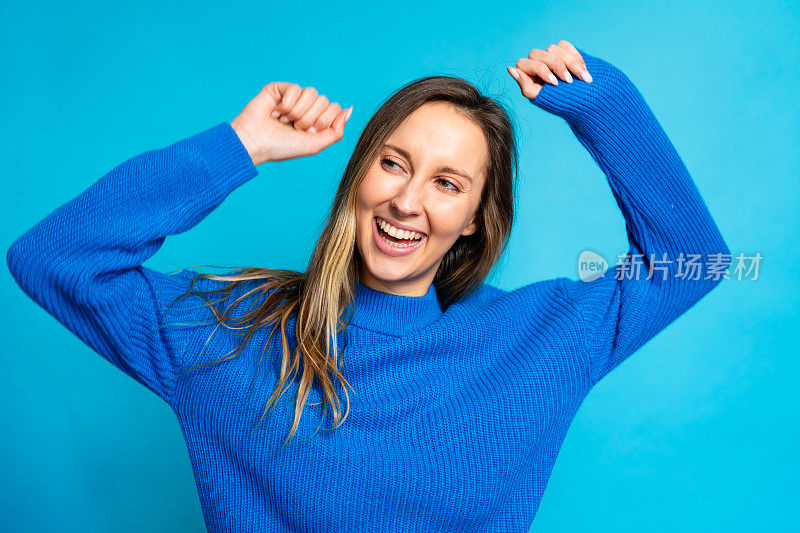 穿着蓝色毛衣的快乐年轻女子在蓝色背景上庆祝胜利