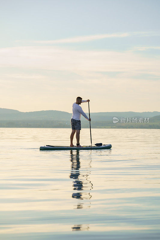 日落时分，一个快乐的人正在湖面上训练冲浪板。站起来划桨。背景是风景。