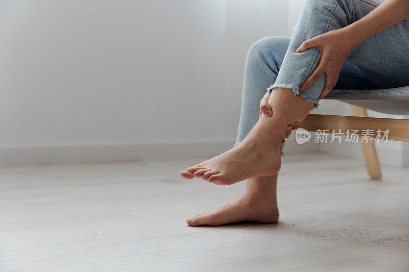 痛苦的腿小腿遭受晒黑美丽的年轻亚洲妇女触摸脚踝在家里室内客厅的特写镜头。伤害，不良健康，疾病概念。酷炫报价横幅