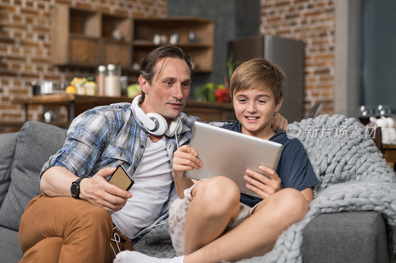 微笑的父亲和儿子坐在沙发上用平板电脑，父母花时间给孩子