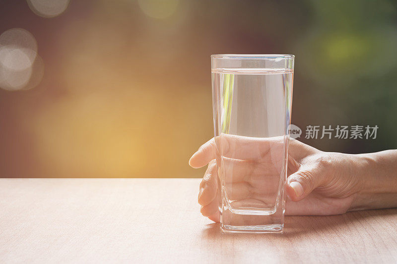 手握一杯纯净水。手拿一杯水放在桌上，以自然为背景。