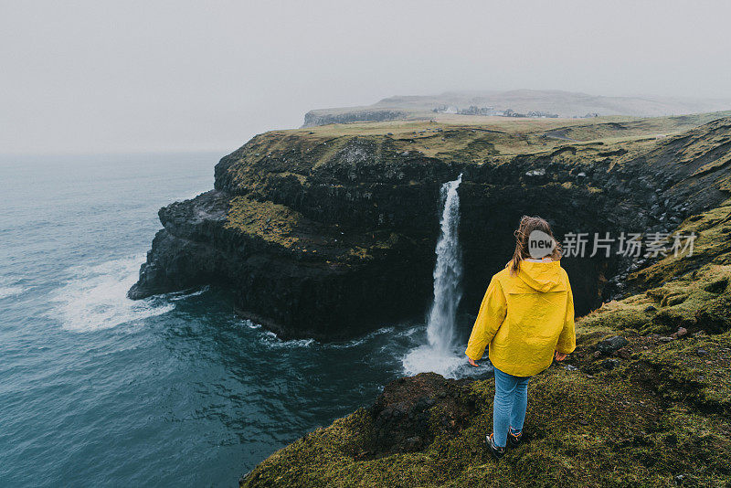 穿着黄色雨衣的女人在看风景Múlafossur瀑布掉进海里在法罗群岛