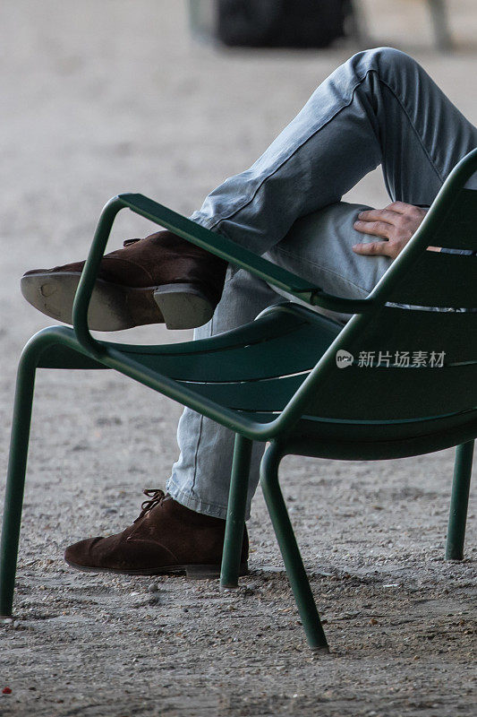 坐在巴黎公园长椅上的男人的双腿和双脚交叉。上等皮鞋和牛仔裤