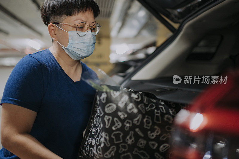 一名戴着口罩的亚裔中国女性外出购物，将东西装进汽车后备箱
