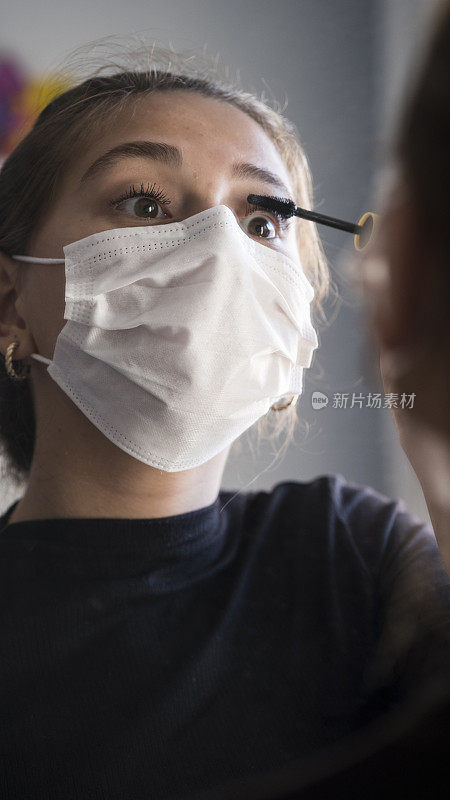 年轻的女人带着医用面具，用睫毛刷化妆，看着镜子