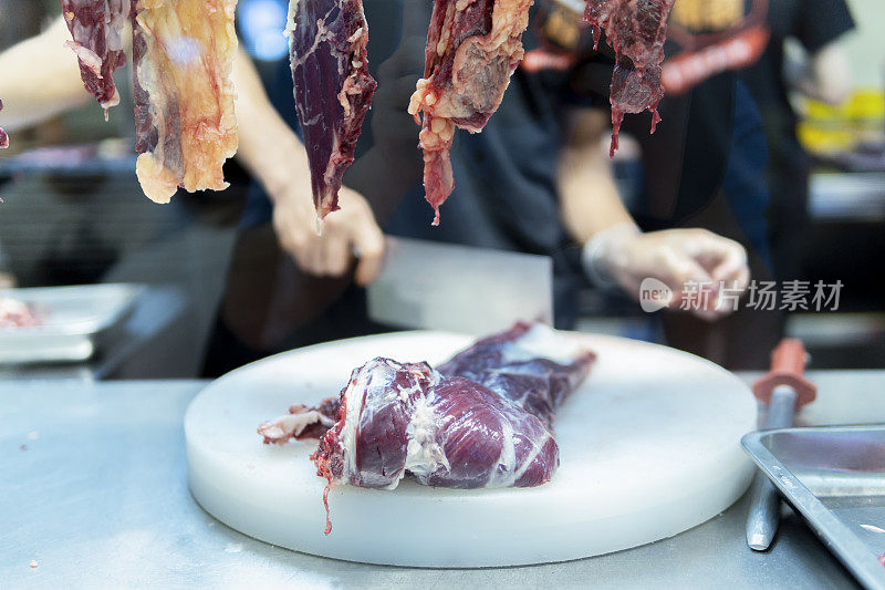 在一家牛肉火锅餐厅的开放式厨房里，厨师在挂着的切碎的生肉后面用刀切牛肉