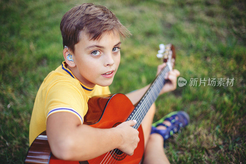 听力不佳的少年在户外弹吉他。带着助听器的孩子在公园里演奏音乐和唱歌。儿童爱好艺术活动。真实的童年的时刻。
