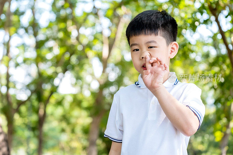 亚洲可爱的男孩或小孩喜欢用手指抠鼻和站在公园里。穿一件白色t恤，上面有复印空间。他试图挤鼻涕。家庭与行为观念。