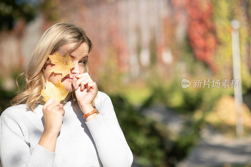 漂亮微笑的中年妇女，金色卷发，卷领毛衣，捧着黄色枫叶近脸。