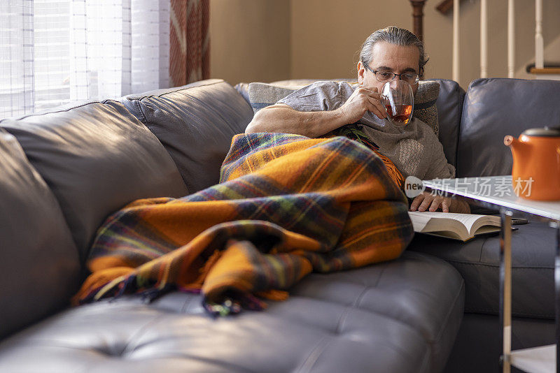 感觉不好。一个成熟的男人生病了，在温暖的格子里躺在沙发上休息。他正在读书，喝着热茶。