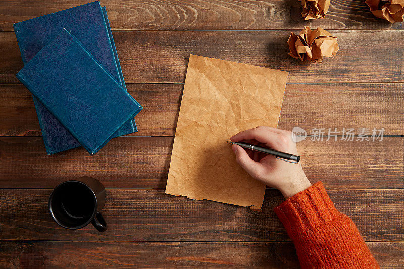 在白纸上写字。木桌上放着一杯咖啡、一本书和皱巴巴的纸。