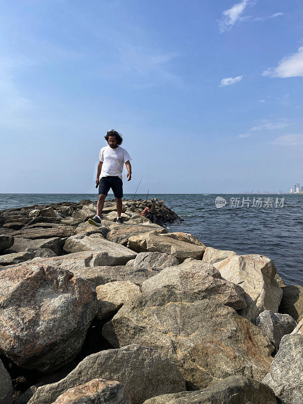 印度人走过斯里兰卡海岸线上人造的岩石海防，海岸侵蚀抛石装甲墙，海岸管理，海洋，背景是斯里兰卡科伦坡的城市摩天大楼