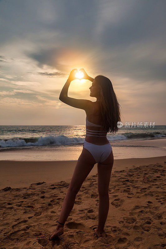 后视图年轻女子做形状的心与手在海边的沙滩日落背景，夏天。在热带海岸享受轻松愉快的假期。旅游度假概念。复制广告文字空间