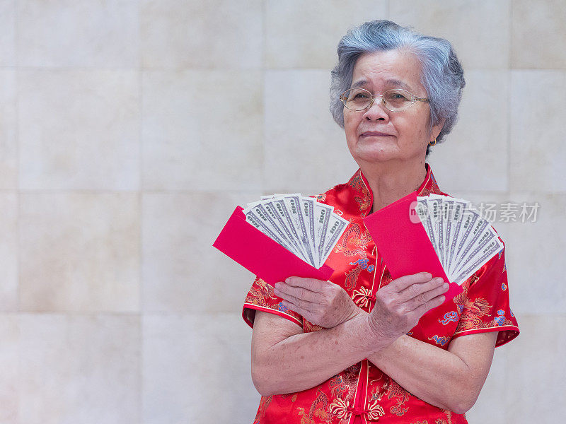 亚洲老妇人穿着旗袍红色衬衫，拿着一个装有美元的红包。