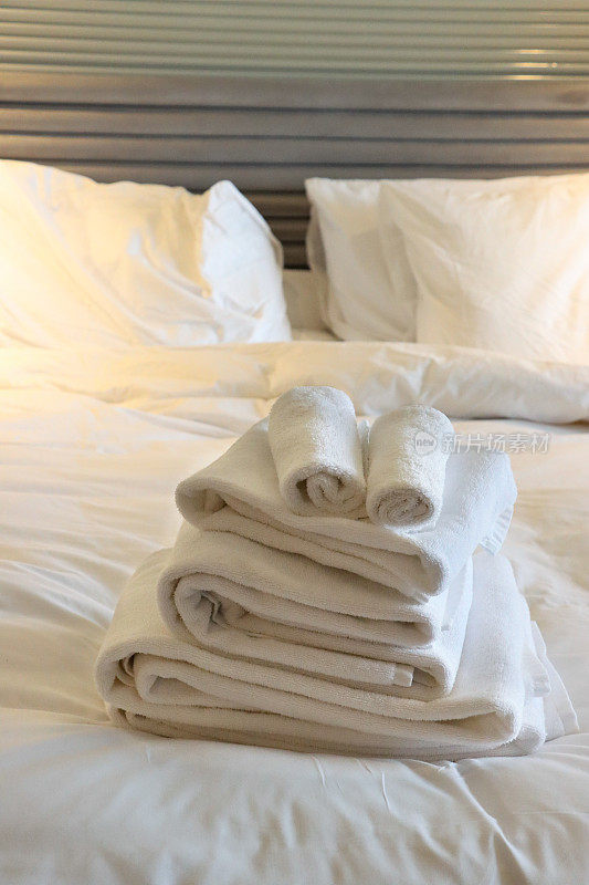 白色酒店毛巾叠在白色羽绒被双人床上，折叠的浴巾和手巾，白色枕头叠在纹理上，指挥棒床头板，重点在前景
